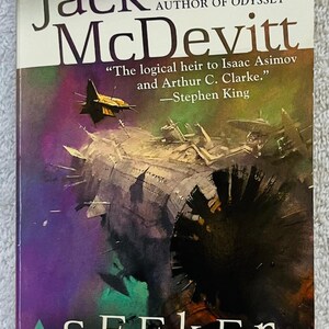 Può includere: Copertina del libro tascabile "Seeker" di Jack McDevitt. L'illustrazione mostra un'astronave in tonalità viola, verde e nero. Il nome dell'autore è in alto, con una citazione di Stephen King. Un romanzo di fantascienza.