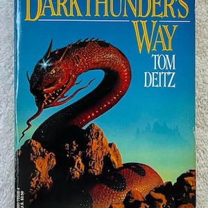 May include: Book cover for "Darkthunder's Way" by Tom Deitz. The cover features a red dragon coiled around a rocky landscape against a blue sky. Text includes the title, author's name, and a tagline.