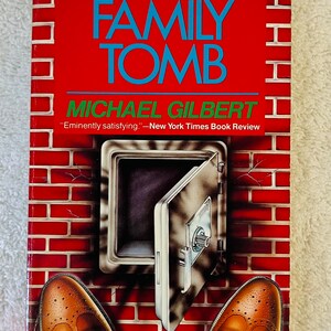 May include: A paperback book titled "The Family Tomb" by Michael Gilbert. The cover features a red brick wall design with a safe door and brown dress shoes. The book has a review quote from the New York Times.