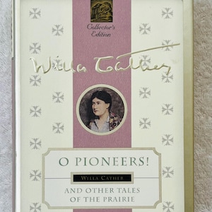 Puede incluir: Un libro de tapa dura titulado "O Pioneers!" de Willa Cather, una edición de coleccionista de The New York Public Library. La portada presenta un retrato del autor, una franja vertical rosa y patrones de cruces decorativos.