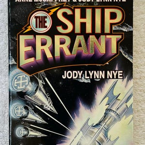 May include: A paperback book titled "The Ship Errant" by Jody Lynn Nye. The cover features a space scene with multiple futuristic ships in shades of silver and white against a dark background. The title is in large, gold-colored letters.