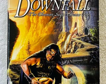 May include: A paperback book titled "Downfall: The Dhamon Saga, Volume One" by Jean Rabe. The cover art depicts a warrior kneeling, holding a pendant, with a fantasy landscape background featuring a castle and bridge. The Dragonlance logo is at the top.