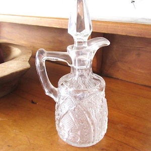 May include: A clear glass cruet with a handle and a stopper. The cruet has a decorative, embossed pattern and a spout. The handle is curved, and the stopper is faceted. The cruet is on a wooden surface.