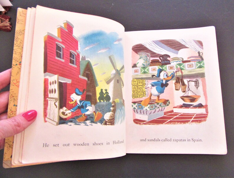 May include: An open book with illustrations of Donald Duck wearing blue sailor clothes. The left page shows Donald Duck in Holland, holding a wooden shoe. The right page shows Donald Duck in Spain, holding a sandal. The text on the left page reads "He set out wooden shoes in Holland". The text on the right page reads "and sandals called zapatas in Spain."