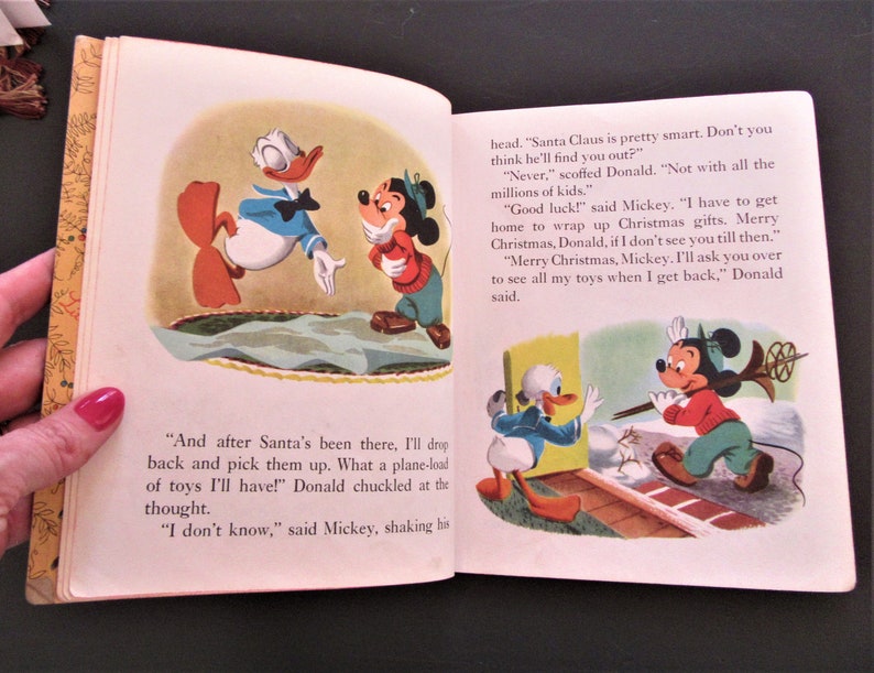 May include: A vintage children's book illustration featuring Mickey Mouse and Donald Duck. Mickey is wearing a red and green sweater and a black hat. Donald is wearing a blue sailor shirt and a red bow tie. The illustration is from a story about Christmas.