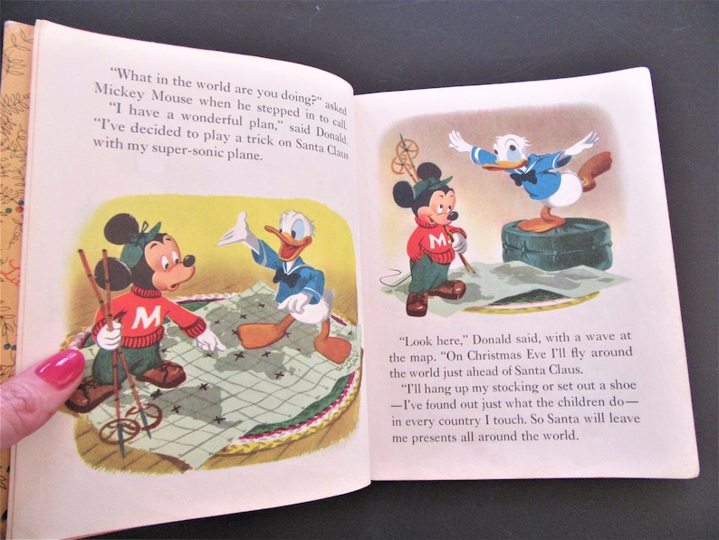 May include: Mickey Mouse and Donald Duck are planning a trick on Santa Claus. Mickey is holding ski poles and looking at a map of the world. Donald is standing on a suitcase and waving at the map. The text reads, "What in the world are you doing?" and "Look here. On Christmas Eve I'll fly around the world just ahead of Santa Claus."