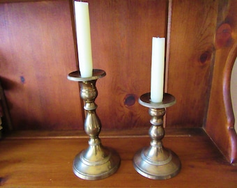 May include: Two brass candle holders with white candles. The candle holders are different sizes and have a classic design.