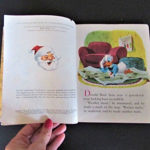May include: An open book with a colorful illustration of Donald Duck drawing on a map. The text reads "Donald Duck bent over a spread-out map, looking busy as could be. 'Wooden shoes,' he murmured, and he made a mark on the map. 'Woolen socks,' he muttered, and he made another mark."