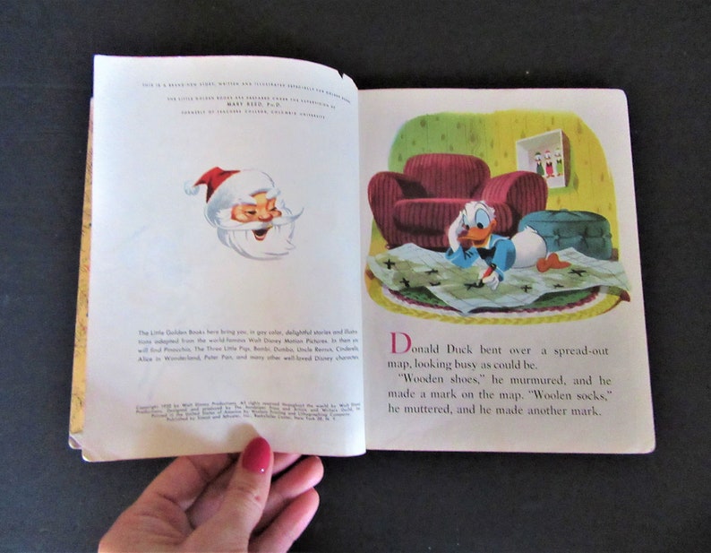 May include: An open book with a colorful illustration of Donald Duck drawing on a map. The text reads "Donald Duck bent over a spread-out map, looking busy as could be. 'Wooden shoes,' he murmured, and he made a mark on the map. 'Woolen socks,' he muttered, and he made another mark."