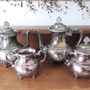 May include: A silver tea set with four pieces: two teapots, a creamer, and a sugar bowl. The set has ornate handles, lids, and decorative floral patterns. The pieces are displayed on a wooden surface.