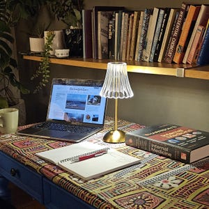May include: A desk setup featuring a silver laptop displaying The New York Times, a gold-toned lamp with a clear fluted shade, a spiral-bound notebook with a red pen, and a thick medical textbook on a desk covered in a colorful geometric patterned fabric.