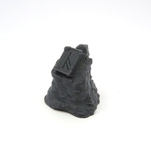 May include: A small, gray, 3D printed rock with a small, gray, rectangular box on top. The box has a raised design on the front.