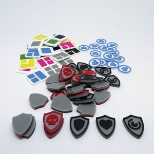 Könnte beinhalten: Eine Sammlung von Spielsteinen, darunter kleine quadratische Kacheln in verschiedenen Farben, runde Token mit blauen und weißen Designs und schildförmige Token mit verschiedenen Symbolen in Schwarz, Rot und Grau.