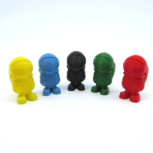 May include: Five small astronaut figurines in a row, each in a different color: yellow, blue, black, green, and red. The figurines are standing upright and have a rounded shape, with a helmet and spacesuit design.