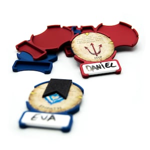 Può includere: Una collezione di pezzi di gioco rossi e blu con gettoni circolari. I gettoni presentano testo e grafica, inclusi i nomi "DANIEL" e "EVA". Un gettone ha la parola "IMP" e un simbolo di ancora. I pezzi del gioco sono su sfondo bianco.