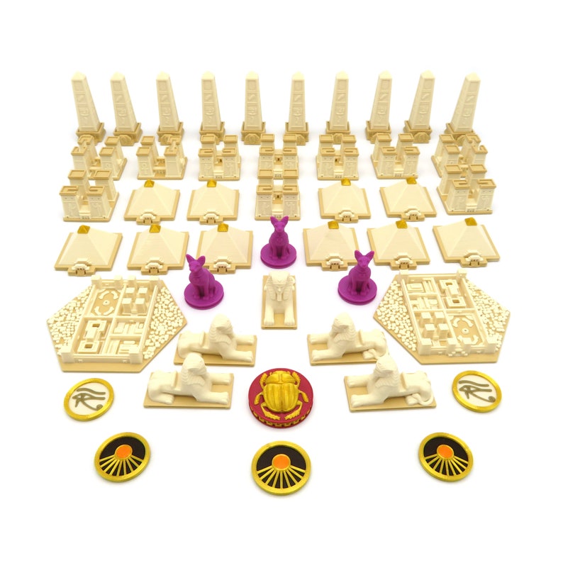May include: A board game set with beige and brown pieces, including small pyramids, obelisks, scarab beetles, and sphinx figures. The game pieces are arranged on a white surface.