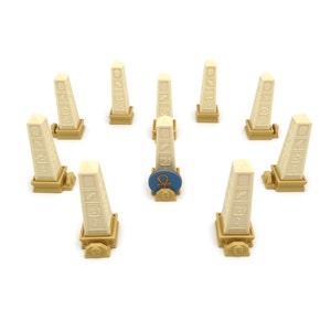 May include: A set of nine beige obelisks with hieroglyphic carvings, each standing on a gold base. One obelisk has a blue and gold circular token with an ankh symbol on top.