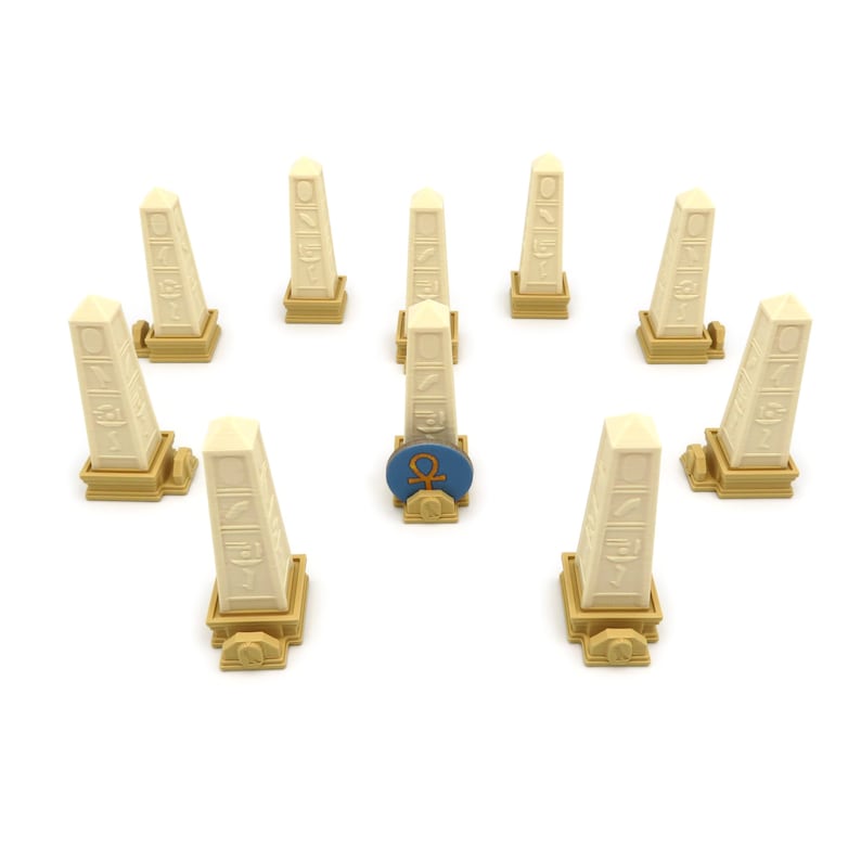 May include: A set of nine beige obelisks with hieroglyphic carvings, each standing on a gold base. One obelisk has a blue and gold circular token with an ankh symbol on top.