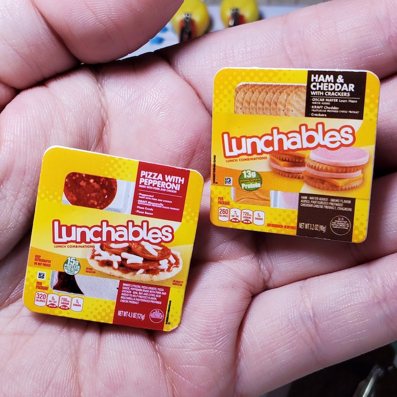 May include: Two Lunchables lunch combinations, one with ham and cheddar crackers and the other with pizza and pepperoni. Both boxes are yellow with red and white accents. The boxes are on a hand.