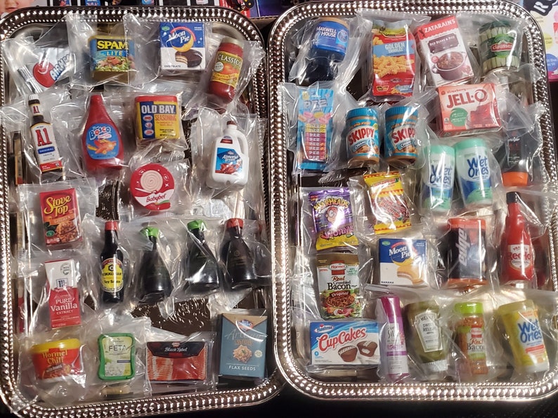 May include: Two silver trays filled with miniature grocery items, including food, condiments, and household products. The items are packaged in clear plastic bags and include brands like Spam, Old Bay, Skippy, Jell-O, and Stove Top.
