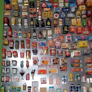 May include: A collection of miniature grocery items, including food, drinks, and household products, displayed on a silver metal surface. The items are arranged in rows and columns, creating a colorful and visually appealing display. Some of the items include: Spam, Jell-O, Cheez-It, Old Bay seasoning, and various brands of soda and juice.
