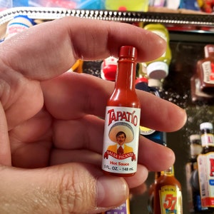 May include: A red glass bottle of Tapatio Salsa Picante hot sauce. The bottle has a white label with a picture of a man wearing a sombrero and the text "Salsa Picante" and "Hot Sauce". The bottle is 5 fluid ounces or 148 milliliters.