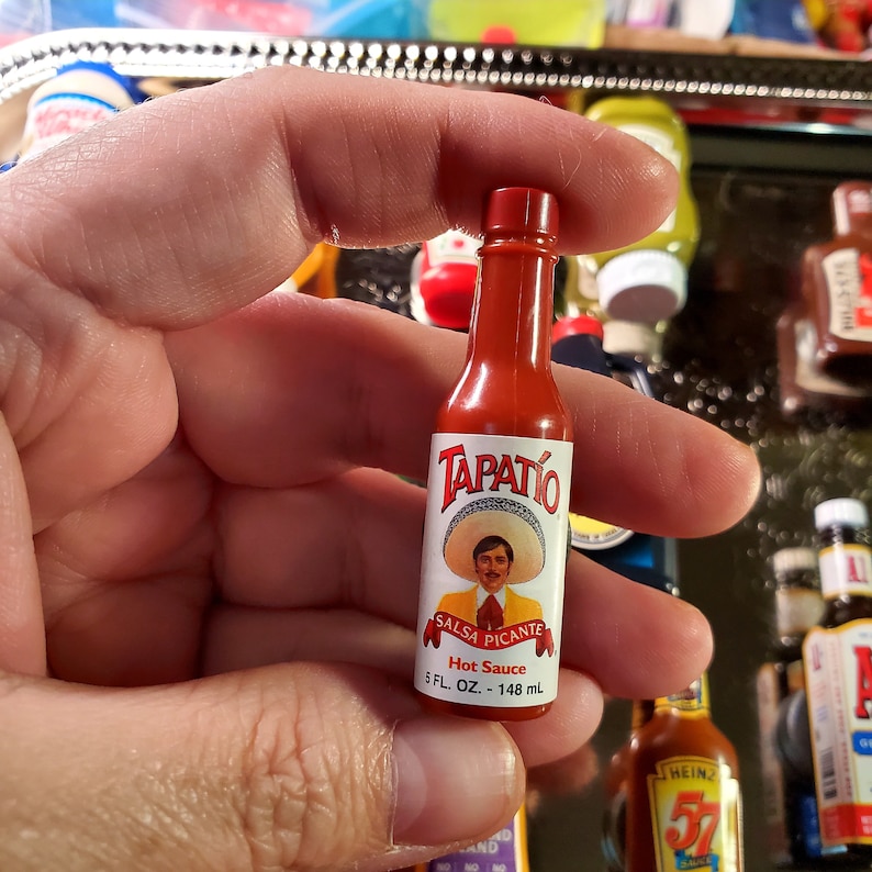 May include: A red glass bottle of Tapatio Salsa Picante hot sauce. The bottle has a white label with a picture of a man wearing a sombrero and the text "Salsa Picante" and "Hot Sauce". The bottle is 5 fluid ounces or 148 milliliters.