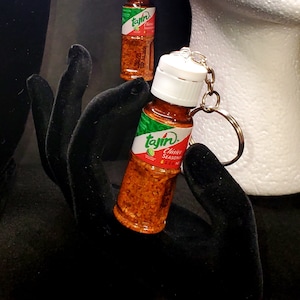 May include: A keychain featuring a miniature bottle of Tajín Clásico Seasoning. The bottle is clear and filled with red and orange seasoning. The bottle has a white lid and a green label with the Tajín logo and text.