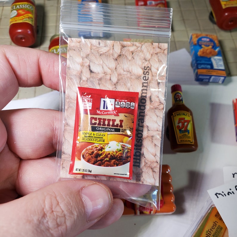 May include: A small plastic bag containing a miniature packet of McCormick Chili seasoning. The packet is red and white with the words "McCormick Chili Original" printed on it. The packet is sitting on a brown and white patterned surface.
