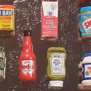 May include: A collection of various food and condiment items, including a jar of dill pickles, a bottle of red hot sauce, a jar of sweet relish, a bottle of mayonnaise, a box of vanilla extract, a jar of peanut butter, and a jar of Old Bay seasoning.