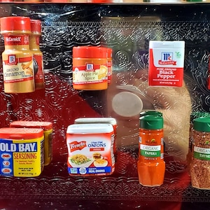 May include: A collection of small spice jars and containers on a silver tray. The jars are red, yellow, and green with white labels. The labels include the names of the spices, such as "Cinnamon", "Apple Pie Spice", "Black Pepper", "Old Bay", "Onions", "Paprika", and "Thyme".