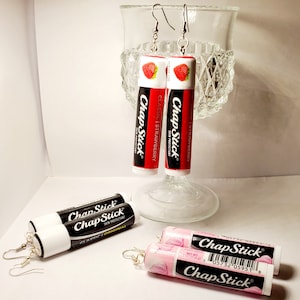 Könnte beinhalten: Ein Paar Ohrringe aus ChapStick Lippenbalsamröhrchen. Die Ohrringe haben die Form der Lippenbalsamröhrchen, mit dem Original-ChapStick-Logo und den Worten "Classic | Strawberry" auf den Röhrchen. Die Ohrringe sind silberfarben und haben einen Hakenverschluss. Es gibt auch ein rosa ChapStick-Röhrchen mit der Aufschrift "ChapStick" darauf.