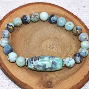 May include: A bracelet made of light blue and dark blue beads, with a central rectangular bead. The bracelet is presented on a wooden surface.