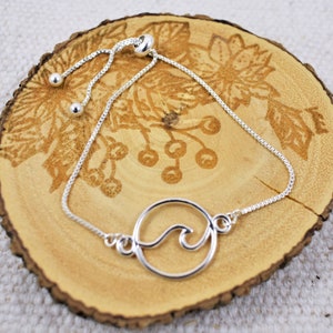 May include: Silver adjustable bracelet with a circle charm featuring a wave design. The bracelet is on a wooden surface.