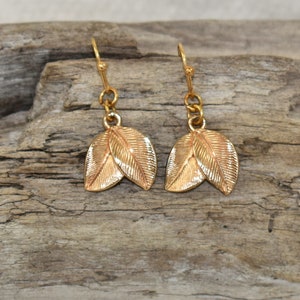 May include: A pair of gold-coloured leaf earrings. Each earring features two detailed leaves, suspended from a simple hook. The earrings are displayed against a textured, light brown wooden surface.