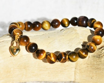 May include: A beaded bracelet featuring tiger's eye gemstones in shades of brown and gold. A gold-colored heart-shaped padlock charm hangs from the bracelet. The bracelet is displayed on a piece of light-colored wood.