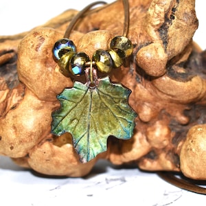 May include: A handmade necklace with a leaf-shaped pendant in green and blue tones. The pendant is adorned with gold-coloured beads and suspended from a brown cord. The necklace is set against a textured wooden backdrop.