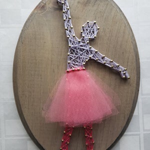 May include: A string art ballerina with a pink tulle skirt on a wooden oval plaque. The ballerina is made with purple string and gold tacks.
