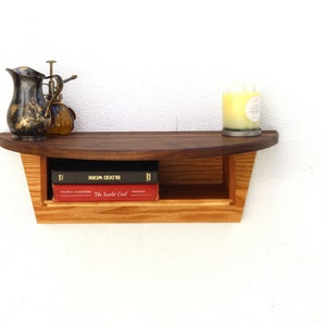 Könnte beinhalten: Ein Wandregal aus Holz mit einer abgerundeten Oberseite und einem unteren Regal. Das Regal besteht aus zwei verschiedenen Holzarten, einer dunklen und einer hellen. Auf dem unteren Regal stehen zwei Bücher, eines rot und eines schwarz. Auf dem oberen Regal stehen eine Kerze und zwei Dekorationsartikel.
