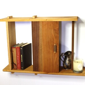 May include: A wooden wall shelf with a small door, two shelves, and a small drawer. The shelf is made of different types of wood, including cherry, walnut, and maple. The shelf is decorated with a small pitcher and a candle.