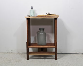 May include: A wooden end table with a shelf and a gray ceramic vase. The table has a light wood top and dark wood sides and legs. There is a book and a bottle of soap on the top shelf.
