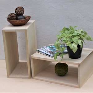 Puede incluir: Dos mesas auxiliares de madera de color claro. Una es alta y rectangular, con una cesta tejida que contiene esferas decorativas en la parte superior. La otra es más baja, con una planta en maceta y revistas. Una esfera verde decorativa se encuentra en el estante inferior.