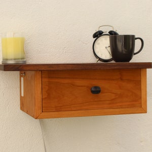 May include: A small wooden wall shelf with a single drawer and a dark brown top. A yellow candle, a black alarm clock, and a black mug are sitting on the top of the shelf.
