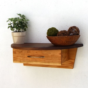May include: A wooden wall shelf with a curved front and a single drawer. The shelf is made of light and dark wood and has a natural finish. The shelf is decorated with a potted plant and a bowl of decorative balls.