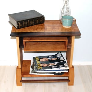 Puede incluir: Una mesa auxiliar de madera con una tapa de color marrón oscuro, un estante de color marrón claro y un estante inferior. La mesa tiene un pequeño cajón en el estante superior. Hay un libro, una taza y una botella de vidrio en el estante superior. Hay revistas en el estante inferior.