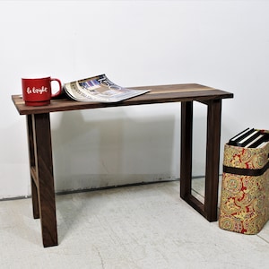 Puede incluir: Un escritorio pequeño de madera oscura con una parte superior rectangular y cuatro patas. Una taza roja con las palabras "be bright" está en el escritorio, junto con una revista abierta. Una caja de almacenamiento estampada está al lado del escritorio.