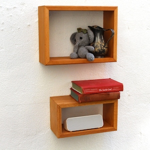 May include: Two wooden wall shelves with a gray elephant stuffed animal, a silver pitcher, and a stack of books on the bottom shelf. The top shelf is a square shape and the bottom shelf is a rectangular shape.