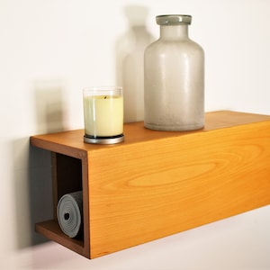 May include: A wooden floating shelf with a natural finish. A rolled-up gray yoga mat is stored in the shelf's open compartment. A lit candle in a glass holder and a frosted glass bottle sit on top, creating a minimalist aesthetic.