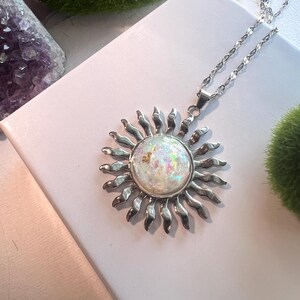 May include: A silver sun pendant necklace with a white iridescent opal stone in the center. The pendant is on a silver chain.
