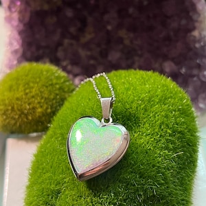Puede incluir: Un colgante en forma de corazón con un centro de ópalo iridiscente y brillante, engastado en un marco de metal plateado. El colgante cuelga de una delicada cadena plateada. El colgante se exhibe sobre musgo verde, con una formación de cristal púrpura en el fondo.